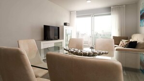 A modern living room with a glass dining table, beige sofas, a flat-screen TV, and a city view through large windows. - Nice Apartment With Shared Pool (Estepona)