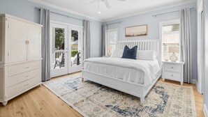 Un dormitorio con una cama blanca, un ventilador de techo, una mesita de noche, un aparador y vistas al exterior a través de un ventanal.