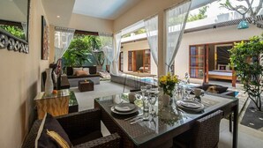 A spacious indoor area with a dining table set for four, surrounded by wicker furniture. There is a seating area with a sofa and a small table, and a view of a swimming pool and outdoor space. - Perenan Villa - Two Bedroom Villa, Sleeps 4 (Pererenan)