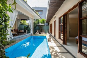 Piscina com deck revestido, área de estar coberta com mesa e cadeiras, e uma estátua no centro.