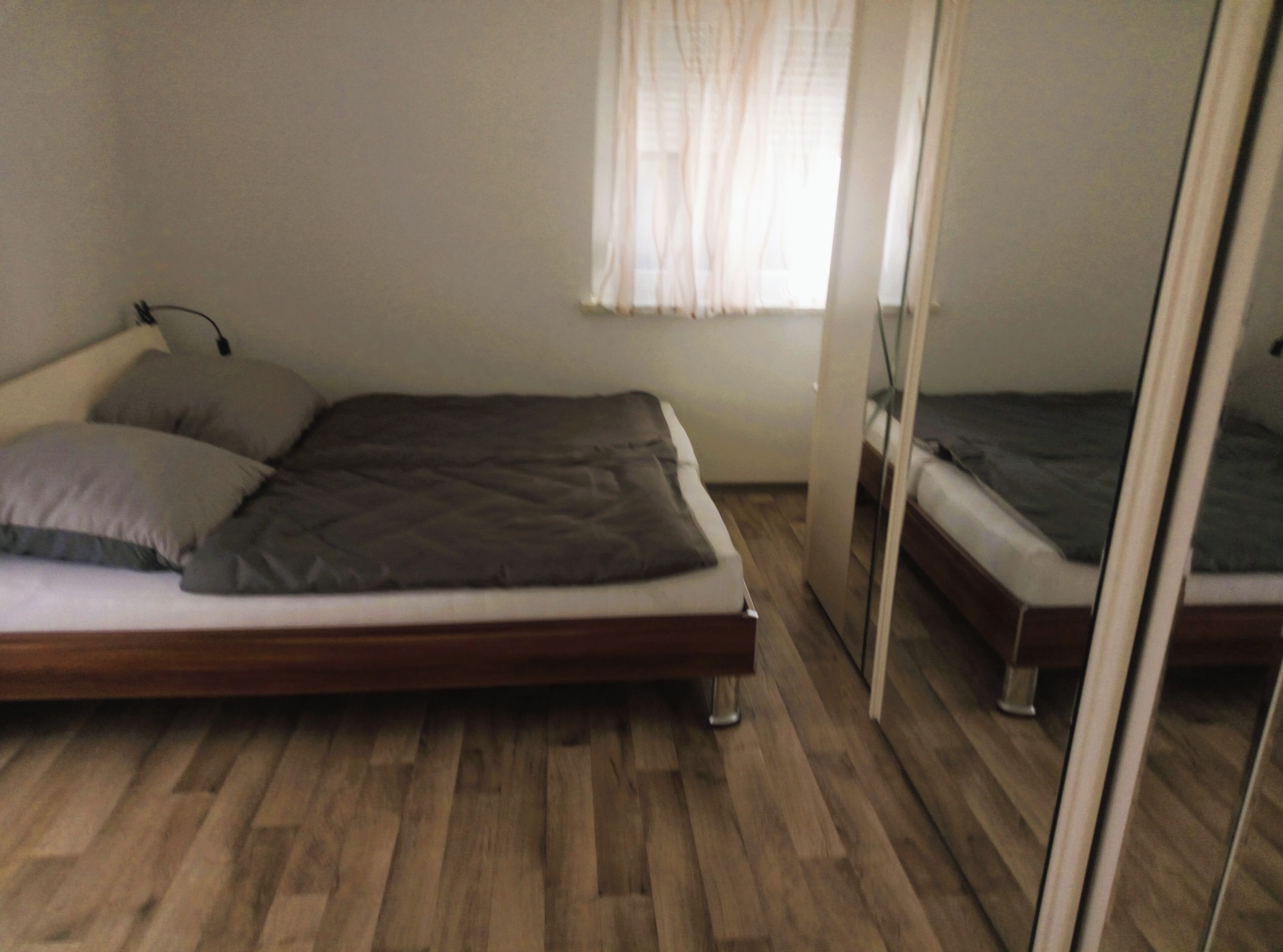 Een eenpersoonsbed met houten frame en matras, een nachtkastje met lamp, en een raam met dunne gordijnen.