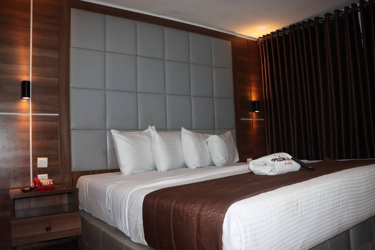 A modern hotel room with a large bed, a headboard with a padded back, a bedside table with a lamp, and a curtain.