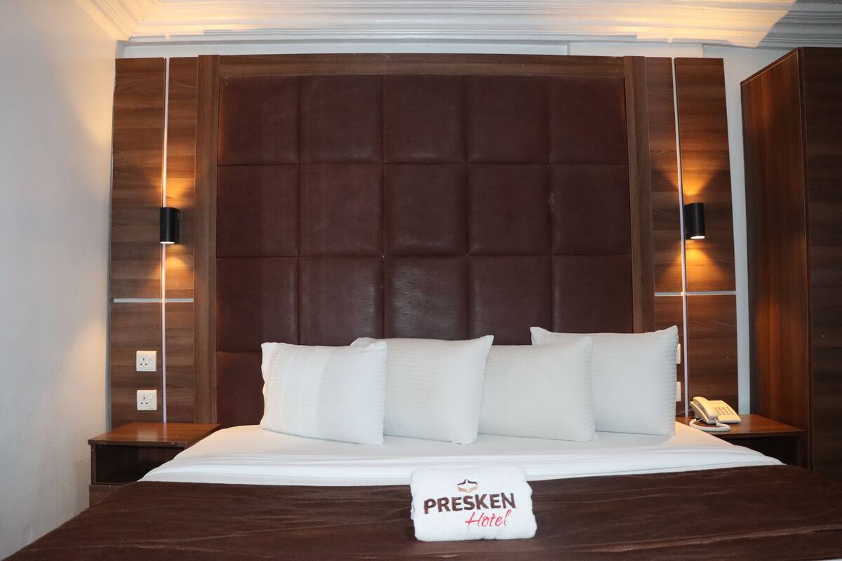 A hotel room with a large bed, two bedside lamps, a nightstand, and a sign reading "PRESKEN Hotel".