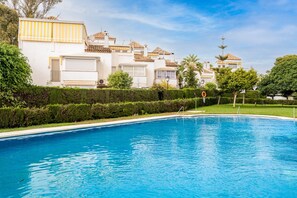 Une piscine à l’eau d’un bleu limpide, entourée de haies soigneusement taillées et bordée d’une rangée de bâtiments blancs aux toits de tuiles.