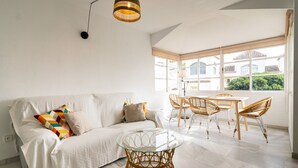 Un salon lumineux avec un canapé blanc, des coussins colorés, une table basse en verre, des fauteuils en osier et une grande fenêtre donnant sur des immeubles et de la verdure.