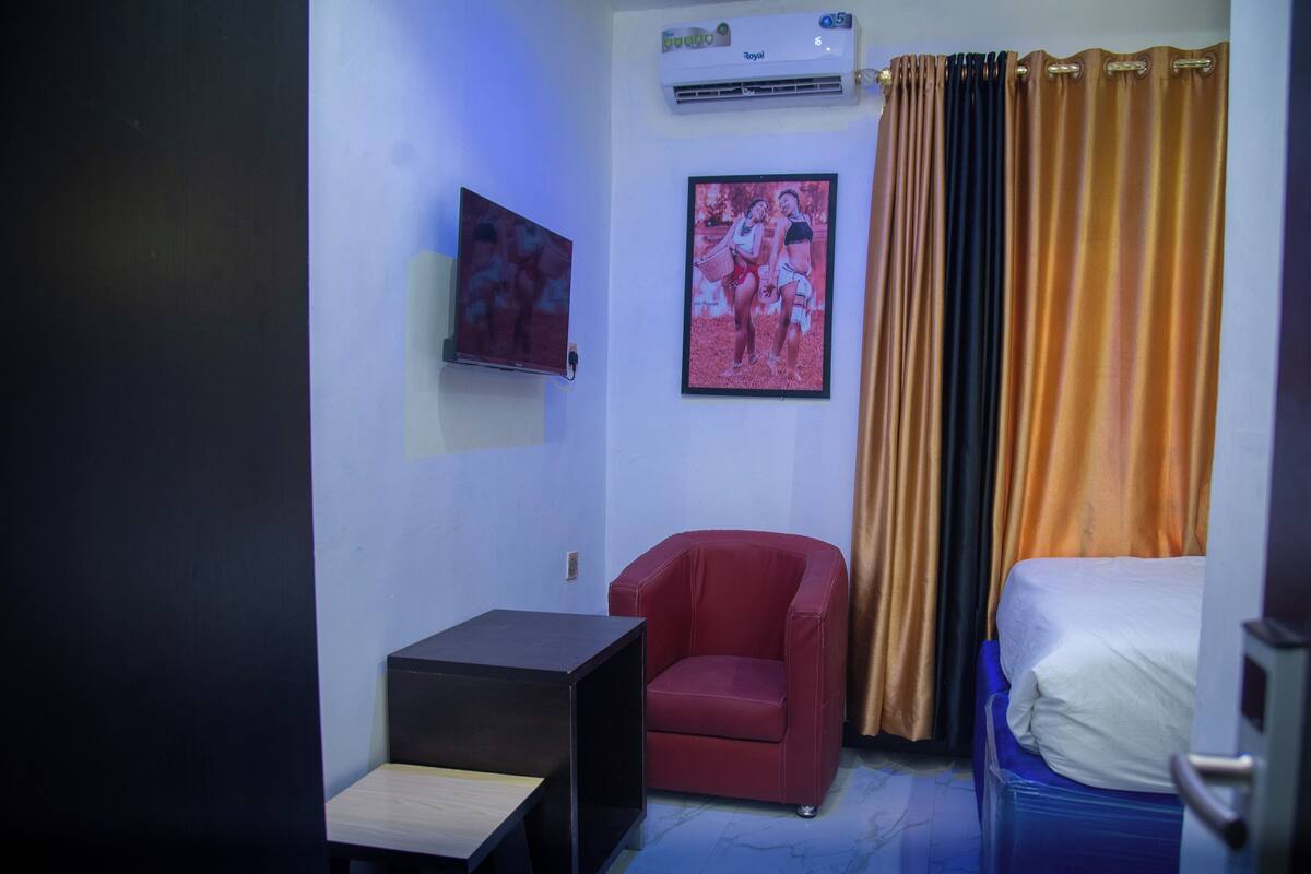Una stanza piccola con un letto, una sedia rossa, un tavolino, una TV a muro e un quadro incorniciato.