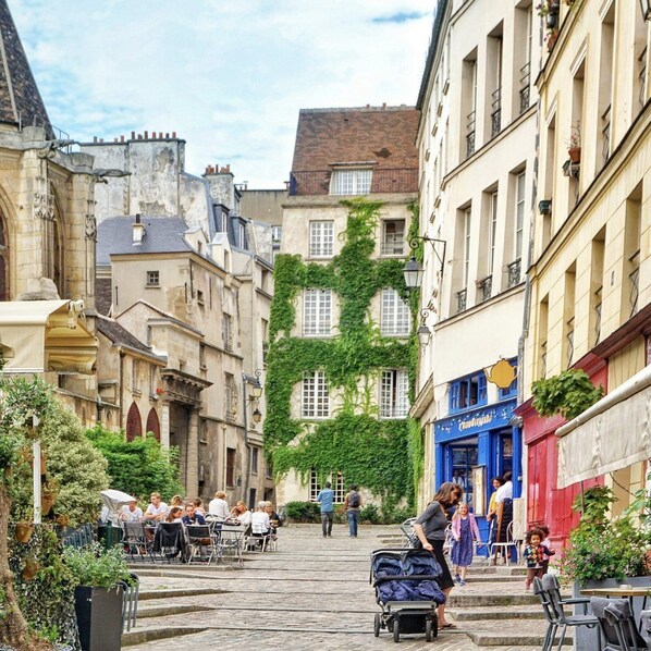 A cobblestone street with buildings on either side, outdoor seating for cafes, and a stroller with a child.