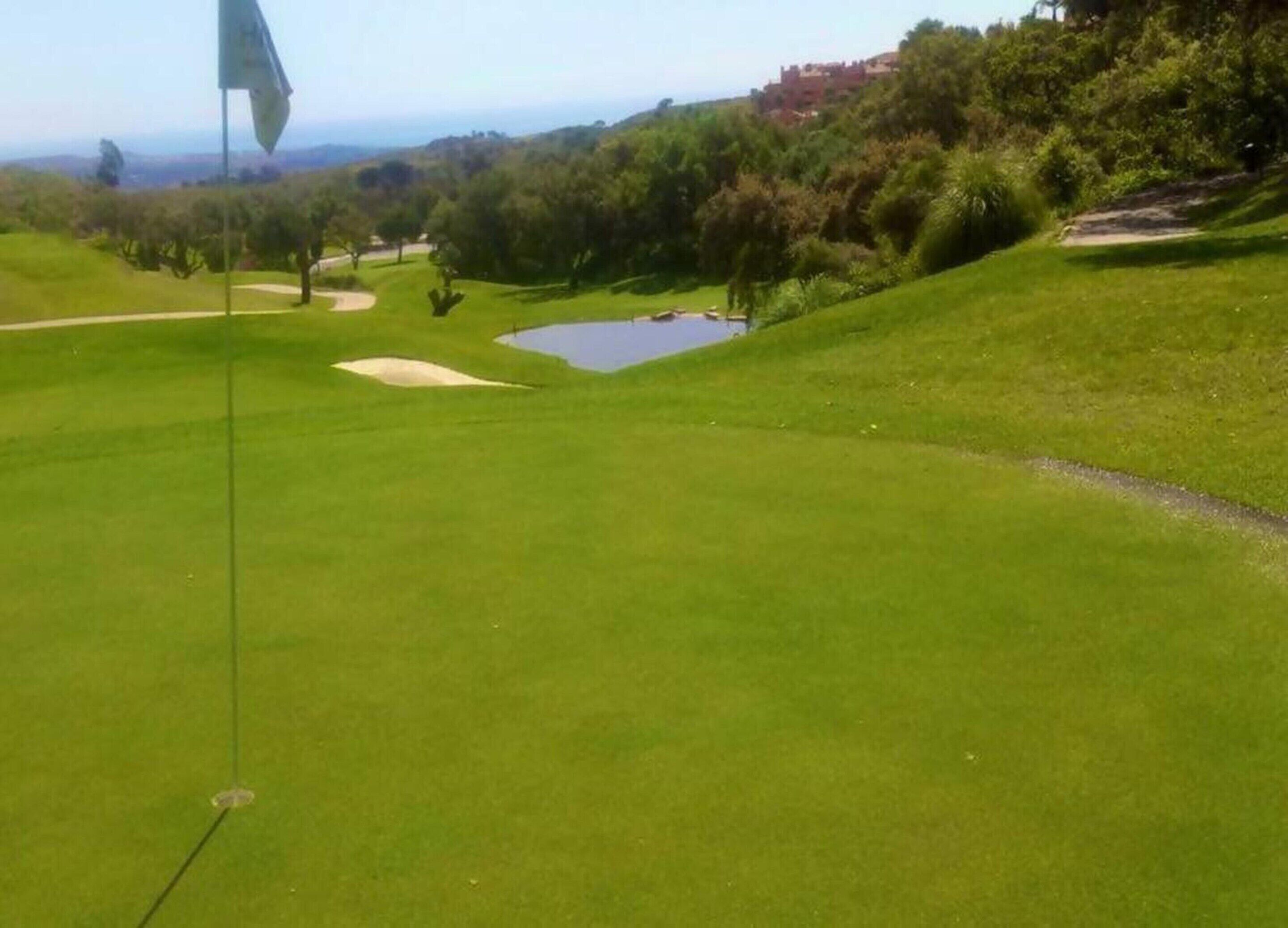 A golf course with a flag, a pond, and a hilly landscape.