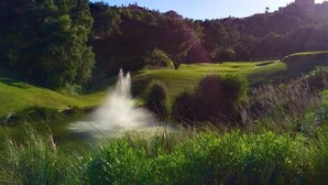 Een golfbaan met een waterfontein, weelderige begroeiing en een heuvel met gebouwen op de achtergrond.