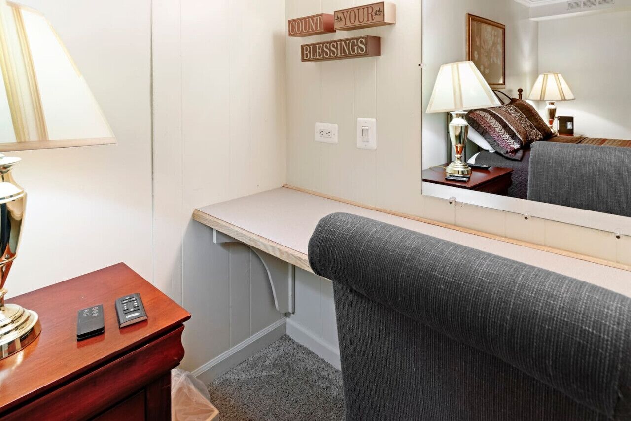 A hotel room with a wooden desk, a lamp, a mirror, and a grey sofa.