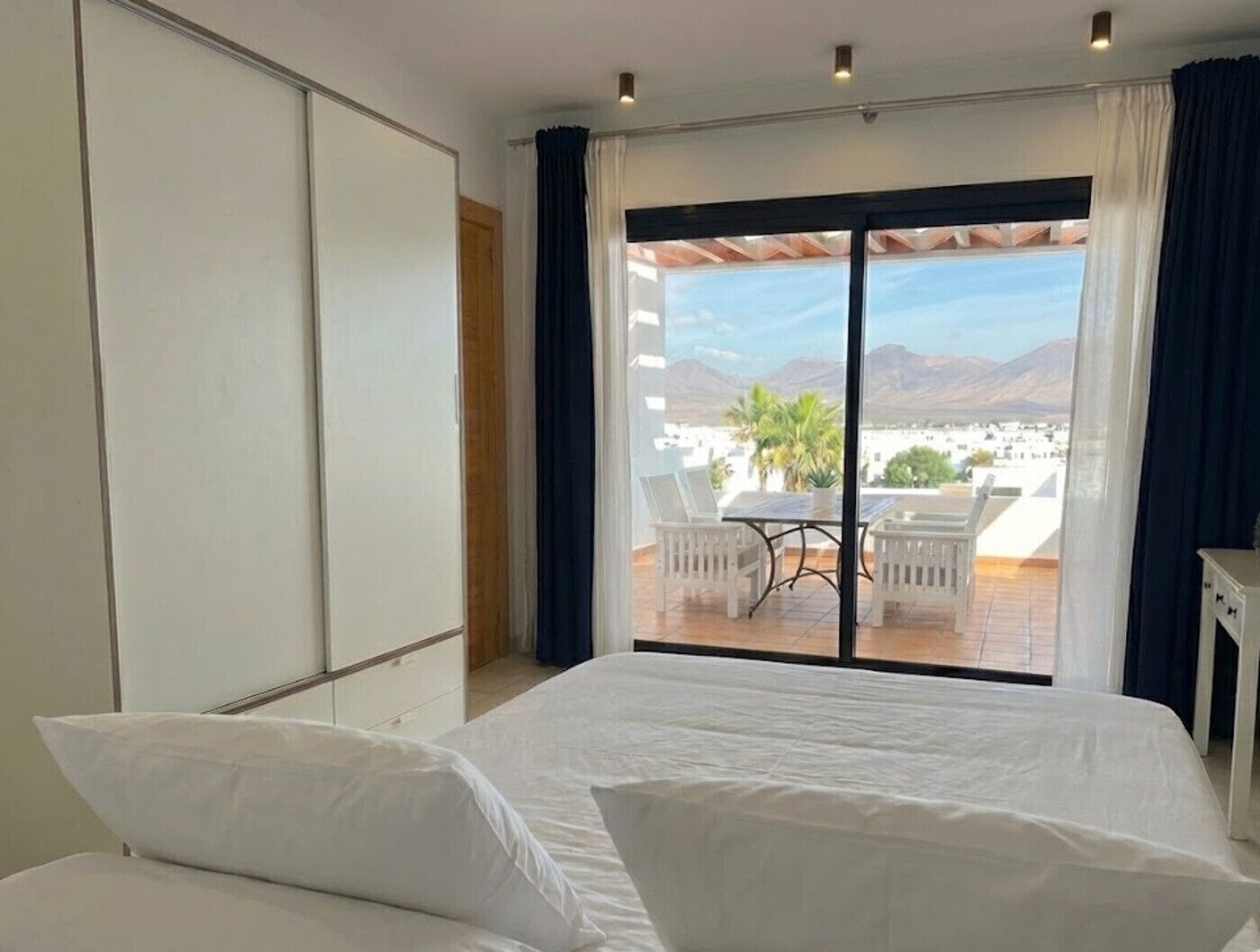 A bedroom with a large bed, a sliding door to a balcony, and a view of the city and mountains.