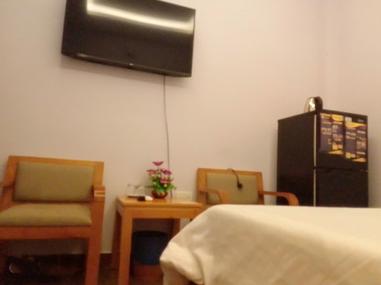 A hotel room with a bed, a desk, two chairs, a flat-screen TV mounted on the wall, and a refrigerator.