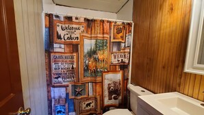 A bathroom with a rustic cabin-themed shower curtain, a white toilet, and a wooden vanity. - The White Stag  (Hillsboro)