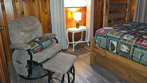 A cozy room with a wooden bunk bed, a rocking chair with a quilt, a small table with a lamp, and a window with blinds. - The White Stag  (Hillsboro)
