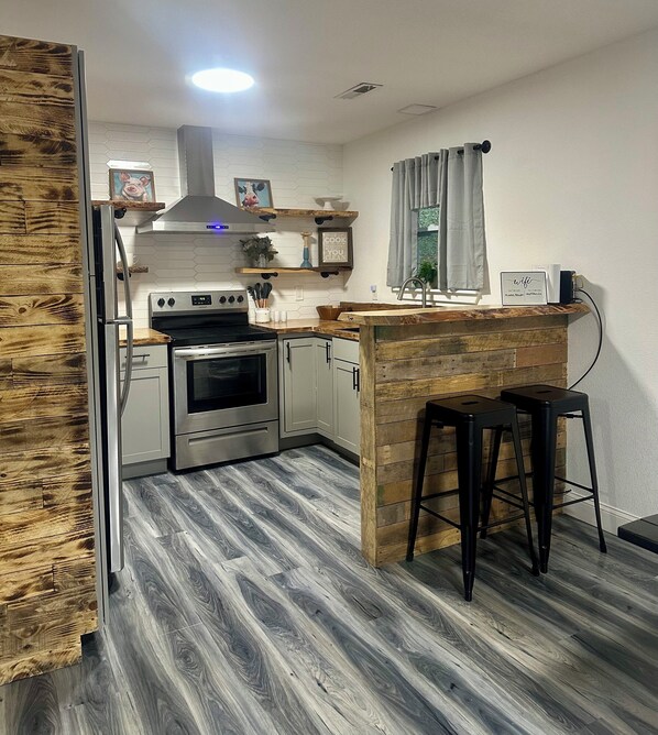 A kitchen with a built-in oven, a stainless steel refrigerator, wooden flooring, and a bar area with stools.