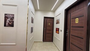 Un pasillo con puerta de madera, pared blanca con cuadros enmarcados y suelo de baldosas.
