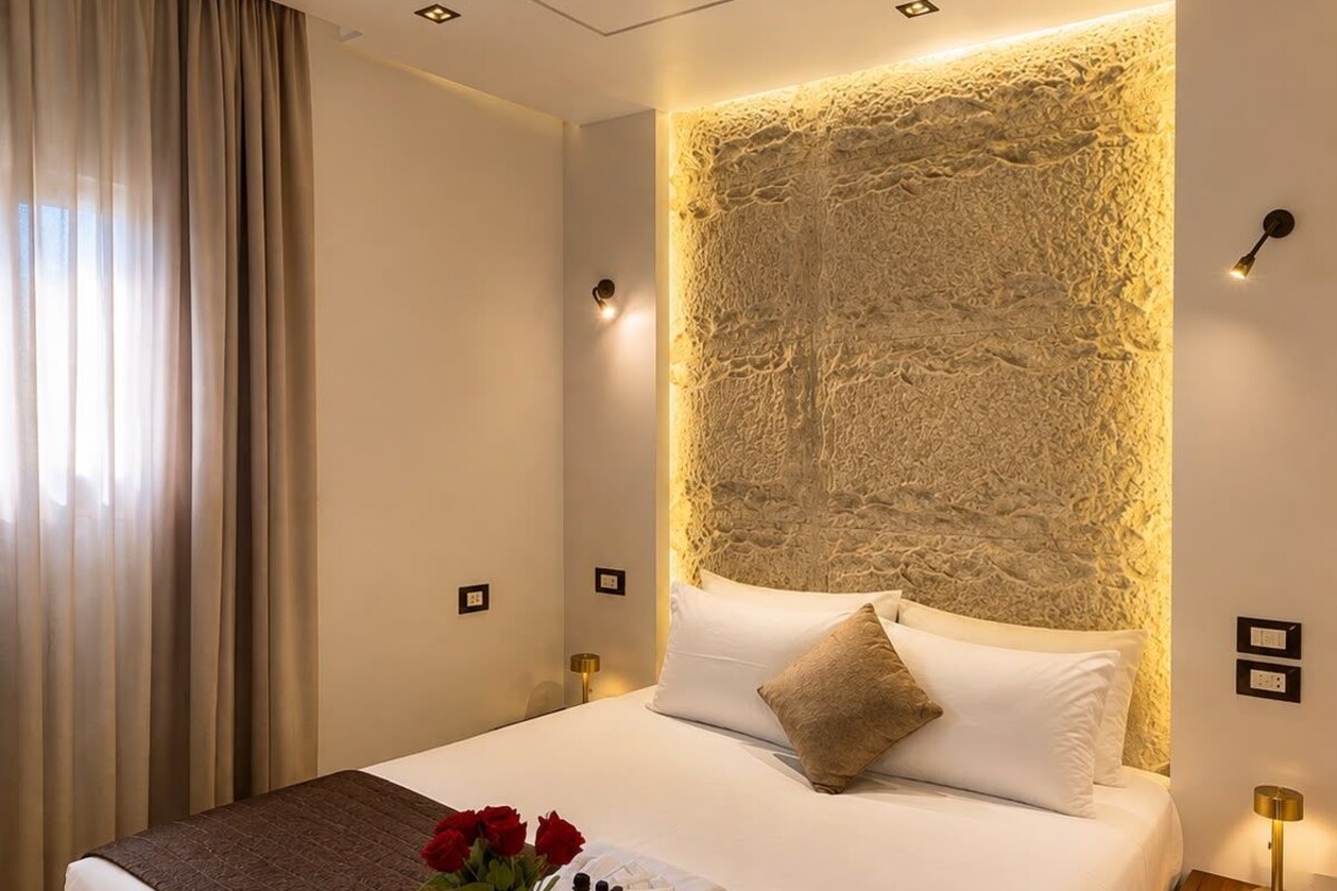 Camera d'albergo con un letto grande, una testata imbottita con decorazione a parete testurizzata, un comodino con lampada e una finestra con tende.