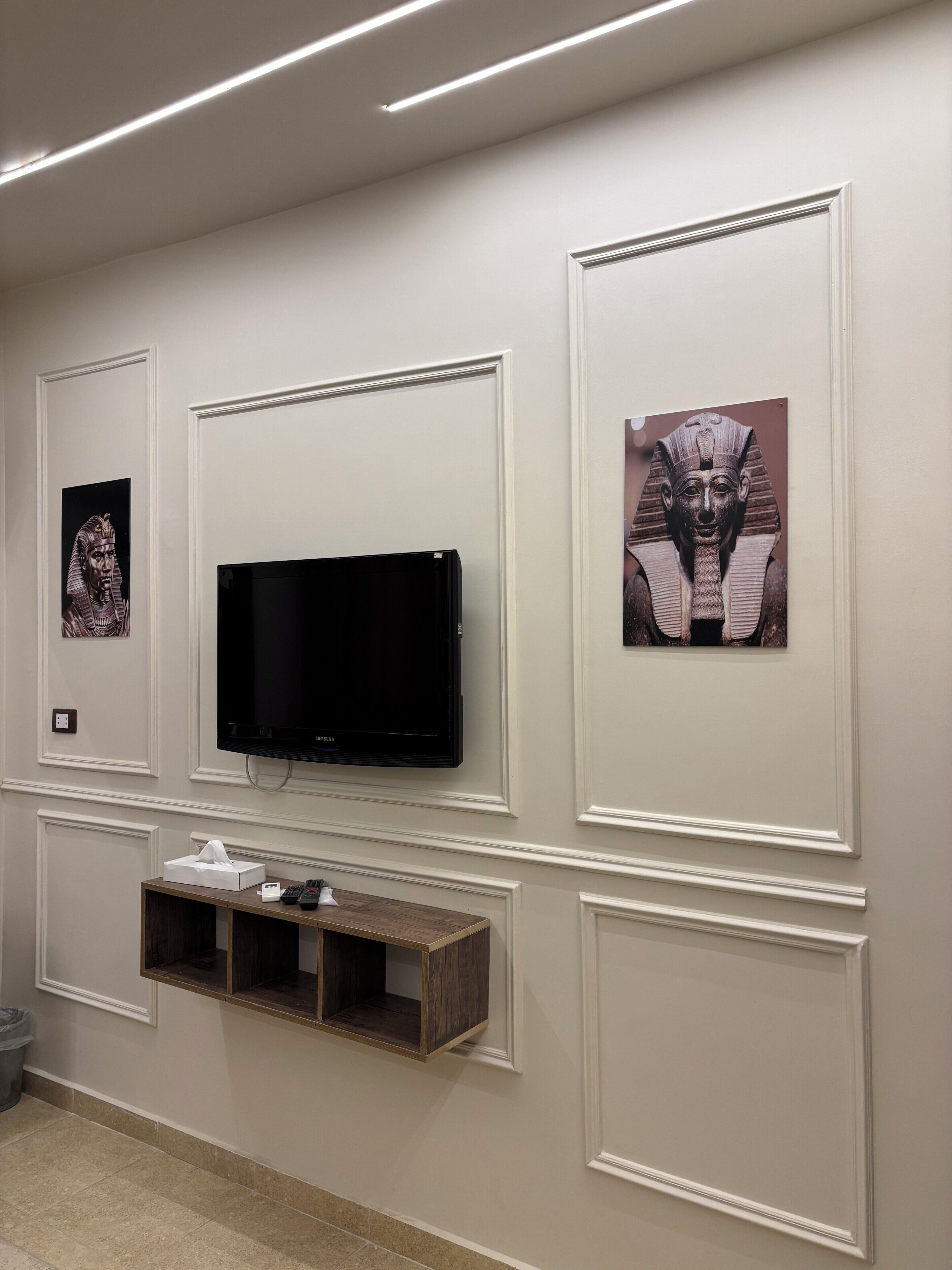 A wall with a flat-screen TV, framed artwork, and a small wooden shelf with a tissue box and remote control.