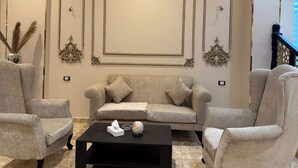 Una sala de estar con un sofá color crema, dos sillones, una mesa de centro negra y molduras decorativas en la pared.
