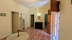 A hallway with patterned flooring, a chandelier, a wooden cabinet, and a door leading to another room. - Cottage Sant Elia - Country Retreat  (Sicilia)