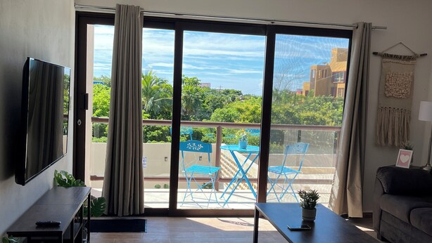 A spacious living room with a flat-screen TV, a sofa, a coffee table, and a balcony with blue chairs and a pool. - Modern&Quiet full Apartment with Pool in Punta Sur, relax and enjoy this place (Isla Mujeres)