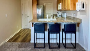 A compact kitchen with a granite countertop, wooden cabinets, a stainless steel refrigerator, and a microwave. There are three bar stools and a small island with a sink. - Spacious 2BR Condo with Secondary Suite on Wolf Creek Golf Course – Sleeps 8! (Mesquite)