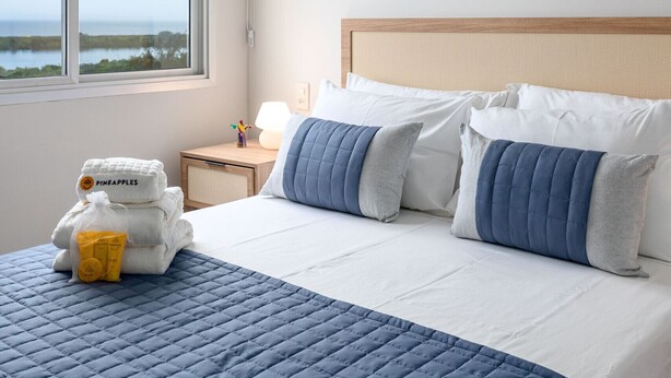 A hotel room with a bed, a nightstand, an air conditioning unit, and a window with a view of the ocean.