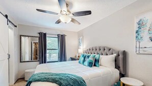 Un dormitorio con una cama grande, un ventilador de techo, una ventana con cortinas, una mesita de noche y una pequeña mesa redonda.