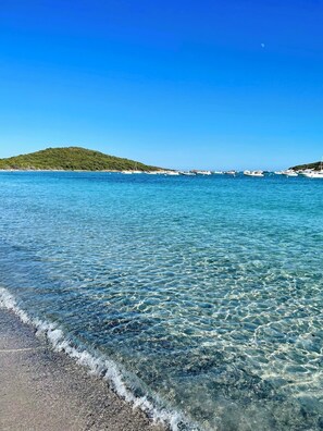 A clear coastal view with calm waters, a sandy beach, and a green hill in the distance.
