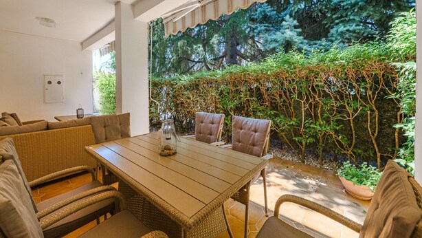 Un comedor con mesa de madera y sillas de mimbre, con vistas a un jardín exuberante con diversas plantas y árboles.