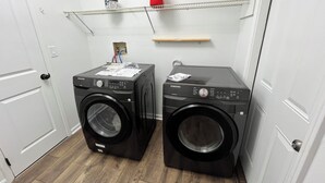 A laundry room with two black Samsung washing machines, a red vase on a shelf, and a wooden shelf above.