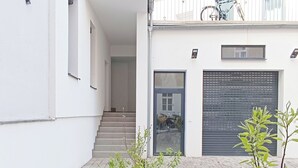 Ein modernes Gebäude mit heller, weißer Fassade, einer überdachten Treppe und einer kleinen Gartenanlage mit jungen Pflanzen.