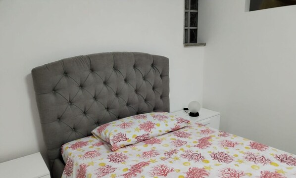 Een slaapkamer met een gestoffeerd hoofdeinde, een bed met een bloemenprint beddegoed, een nachtkastje met een lamp en een klein raam.