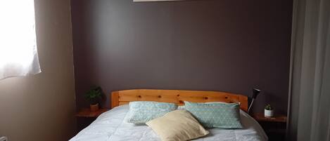 Een netjes opgemaakt bed met een houten hoofdbord, een ingelijst kunstwerk erboven en een klein plantje op het nachtkastje.