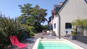 A swimming pool with a wooden deck, a red lounge chair, and a house with a gabled roof. - Charming country house with swimming pool in Vannes for family vacations (Vannes)