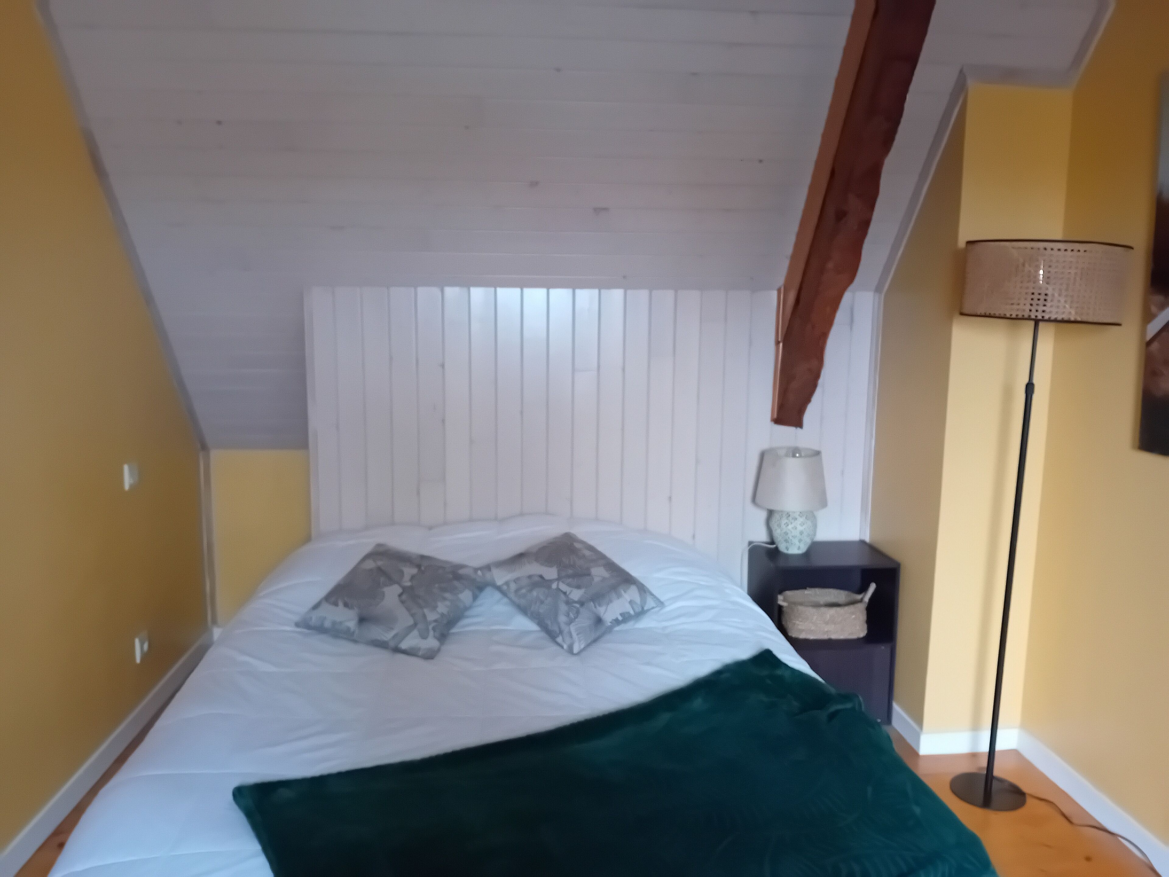 Een slaapkamer met een schuine kap, een bed met witte beddengoed en twee kussens, een groene plaid, een nachtkastje met een lamp, en een vloerlamp.