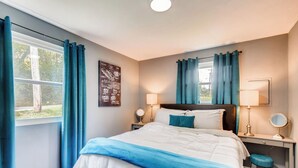 Un dormitorio con cama, dos mesitas de noche con lámparas, un escritorio con silla y una ventana con cortinas.