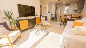 Een moderne woonkamer met een bank, fauteuils, een salontafel en een televisie. Achtergrond toont een keuken met een eettafel en stoelen.