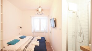 A bedroom with a bed, a bedside table, a window with curtains, a shower area with a glass door, and a hanging light fixture. - Zaratino rooms (Zadar)