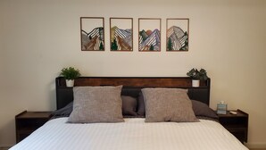 Un llit ben fet amb edredó blanc i gris amb ratlles, dos coixins a joc i un panell frontal de fusta. Hi ha dues taules de nit amb petites plantes en testos i una làmpada de nit. Sobre el llit, hi ha quatre quadres amb paisatges abstractes de muntanyes.