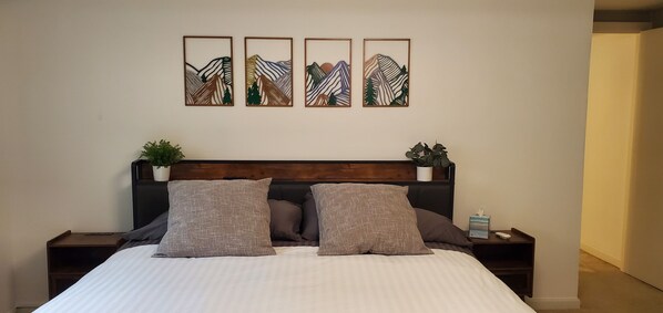 A neatly made bed with a white and gray striped duvet, two matching pillows, and a wooden headboard. There are two bedside tables with small potted plants and a bedside lamp. Above the bed, there are four framed artworks depicting abstract mountain landscapes.