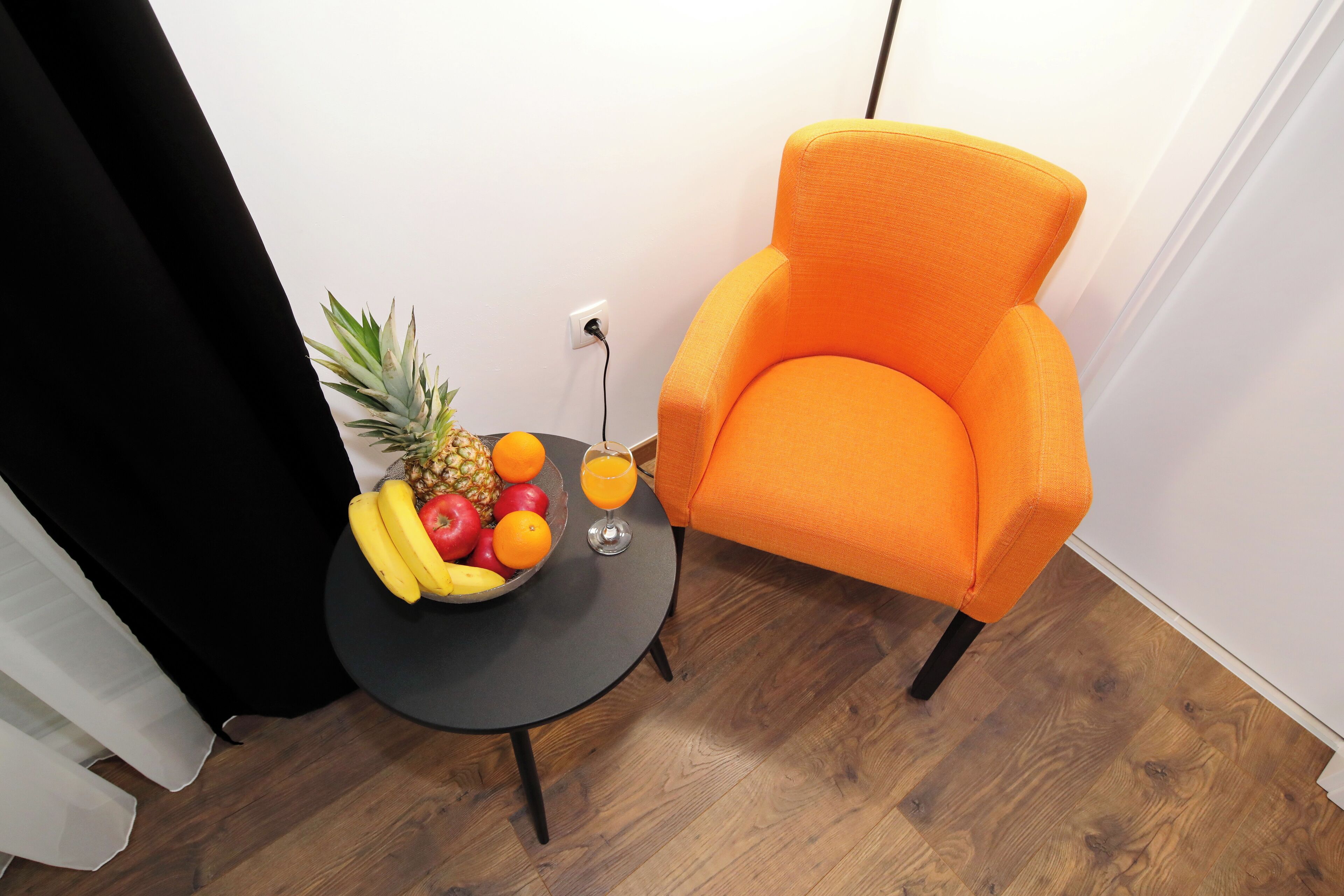 A room with a wooden floor, a black table with a fruit bowl and a glass of orange juice, and an orange armchair.