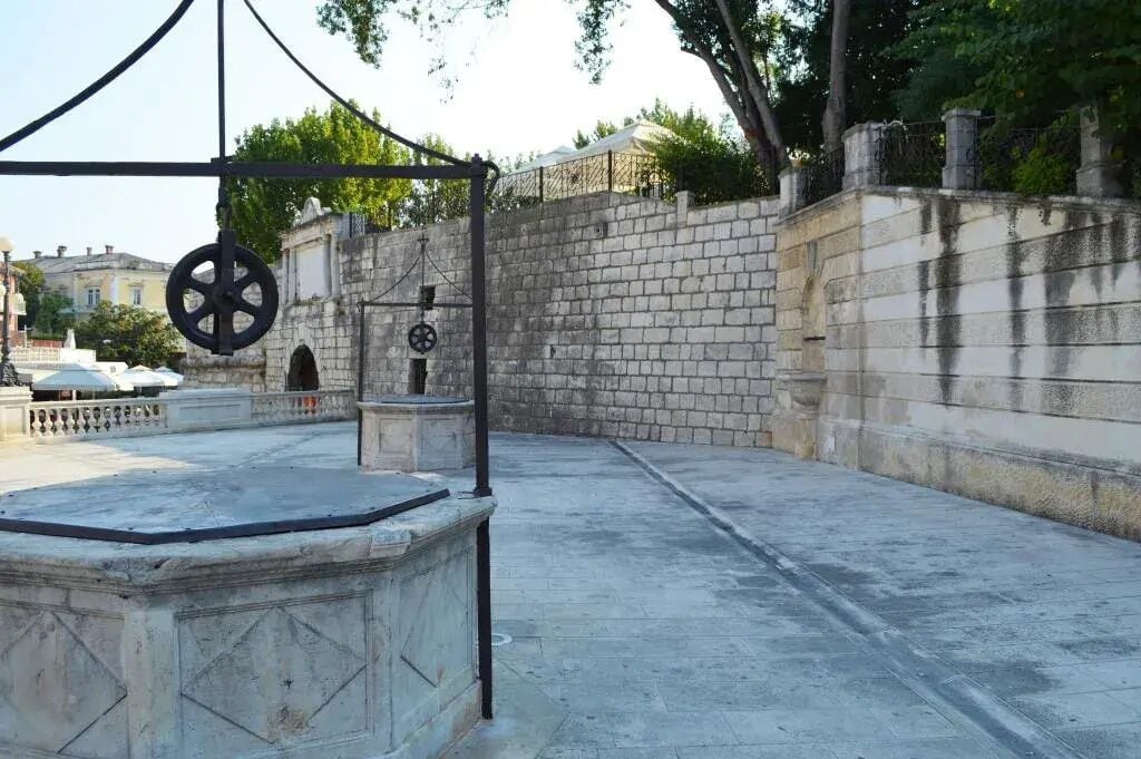 A courtyard with a stone wall, a fountain, and a metal structure with wheels.
