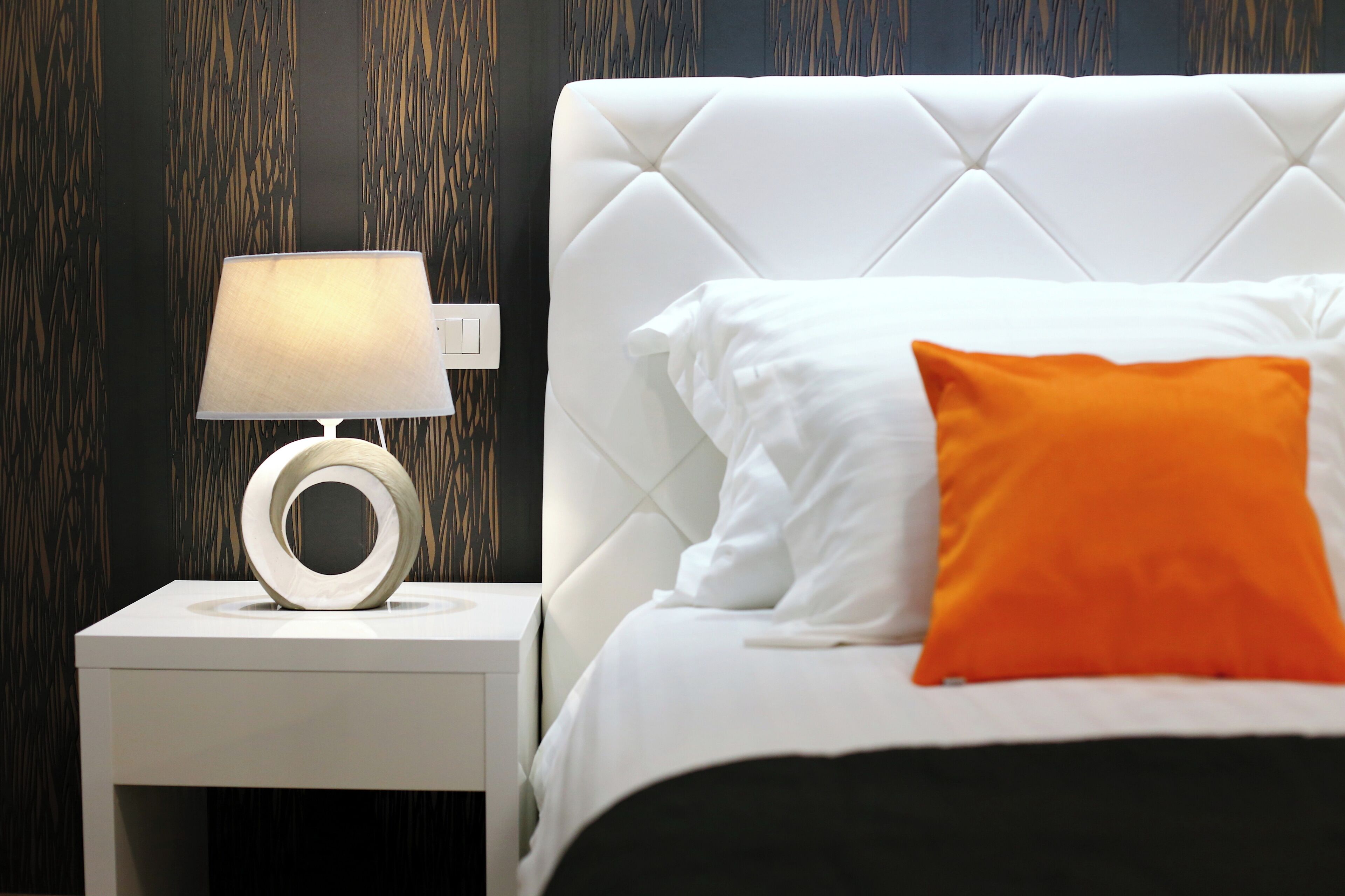 Una cama bien hecha con almohadas blancas y anaranjadas, una mesita de noche blanca con una lámpara moderna y una pared oscura con estampado.