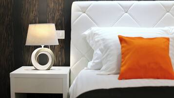 Una cama bien hecha con almohadas blancas y anaranjadas, una mesita de noche blanca con una lámpara moderna y una pared oscura con estampado.