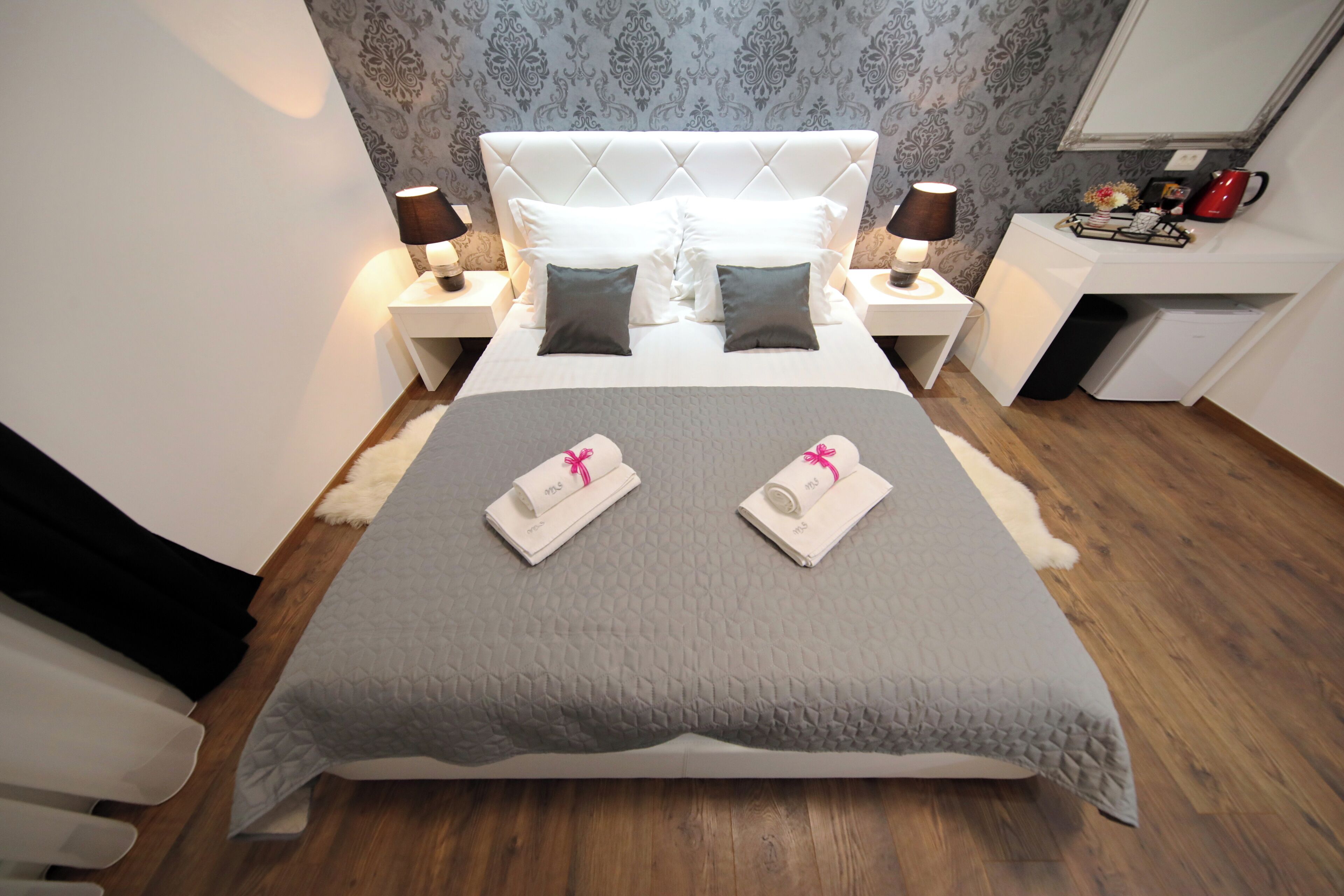 A neatly made bed with a grey quilt, two white pillows, and two rolled towels with pink ribbons. There are two bedside tables with lamps, a small shelf with a red kettle, and a white cabinet. The room has patterned wallpaper and wooden flooring.