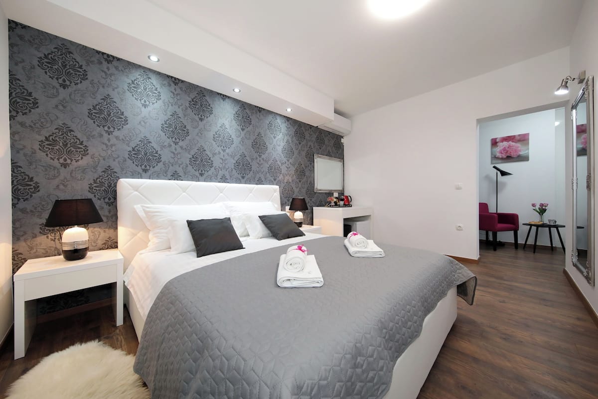 Ein modernes Hotelzimmer mit einem großen Bett, einem Nachttisch mit Lampe, einem kleinen Tisch ebenfalls mit Lampe, einem Sessel und einem Spiegel.