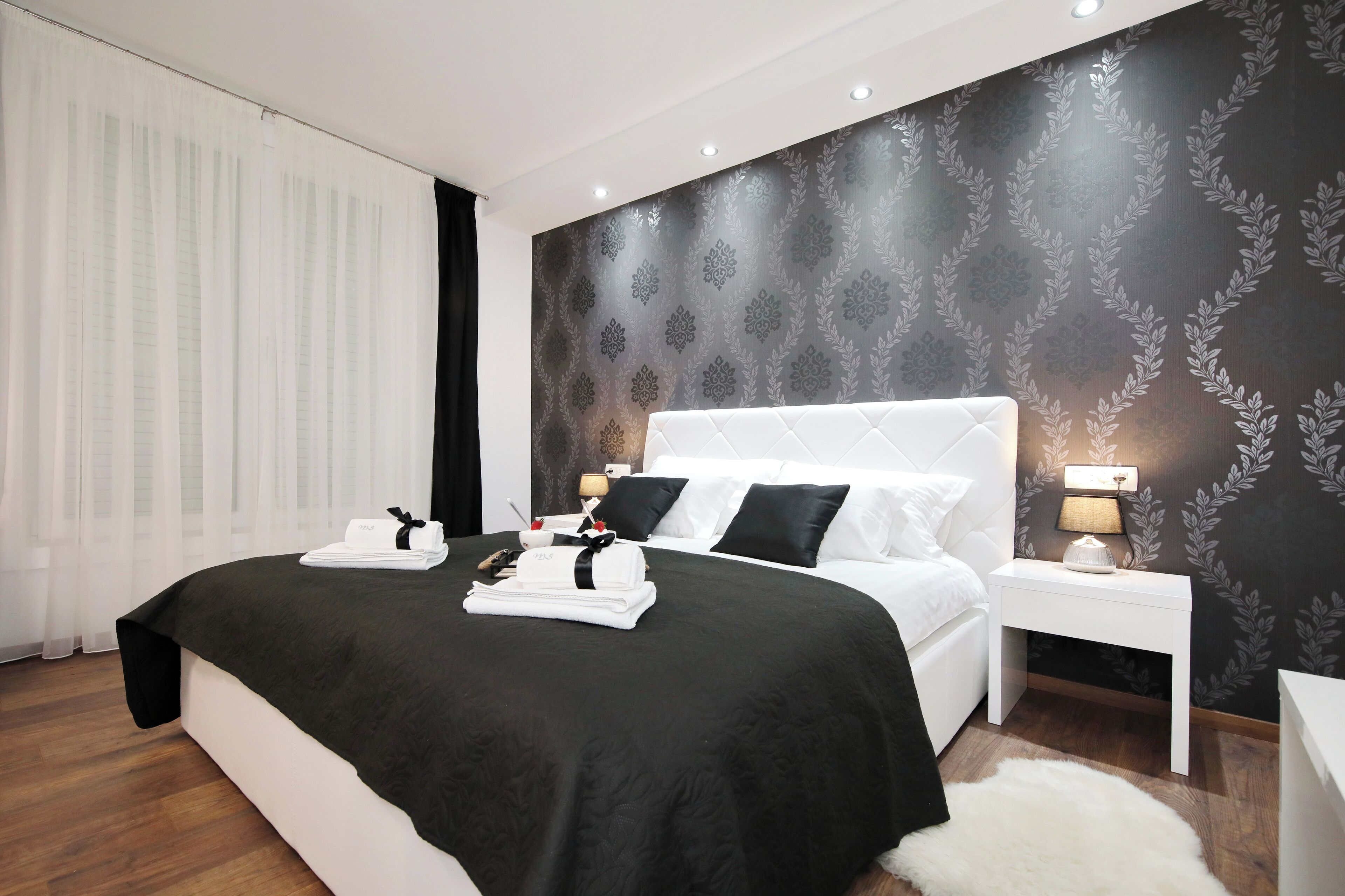 A modern hotel room with a large bed, white bedding, a dark patterned wall, and a small bedside table with a lamp.