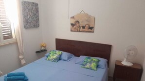 A neatly made bed with blue linens and pillows, a bedside table with a fan, and a wall with a floral painting.
