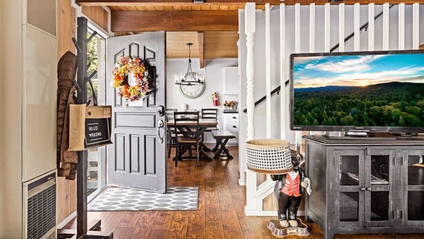 A cozy living room with a wooden ceiling, a TV on a cabinet, a dining area with a chandelier, and a decorative wreath on the door. - Guest FAV Luxury Sun Chalet + Hot Tub + BBQ (Big Bear Lake)